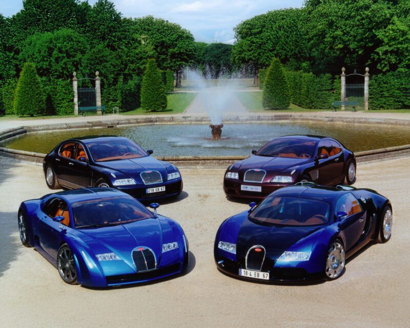 Four Bugatti cars are parked in front of a round fountain in a landscaped garden, with trees and a gate in the background.