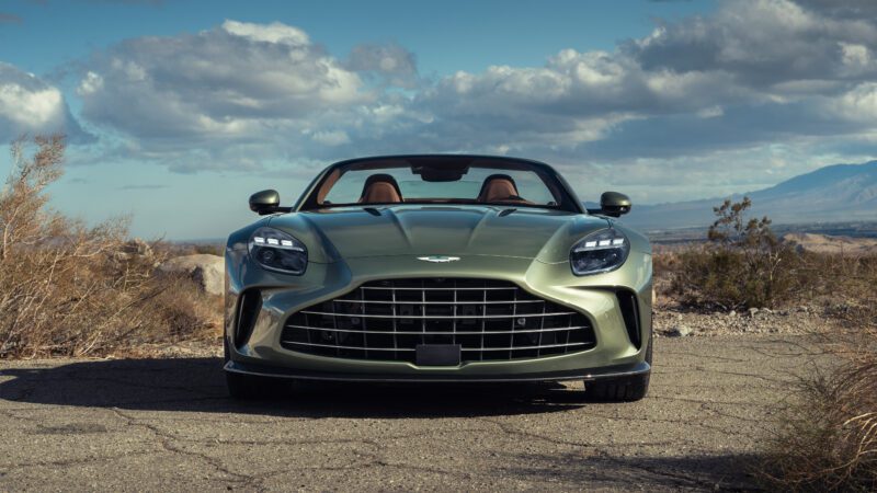 A green convertible sports car with a large front grille is parked on a cracked road in a desert landscape under a partly cloudy sky.