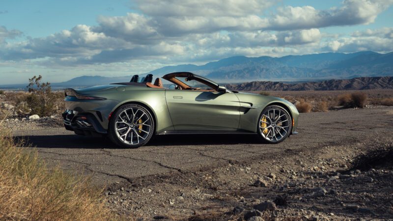 A green convertible sports car parked on a cracked asphalt road in a desert landscape with mountains in the background under a partly cloudy sky.