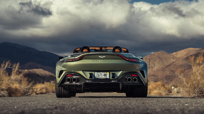 A green convertible sports car, seen from the rear, is parked on a deserted road surrounded by dry grass and mountains under a cloudy sky.