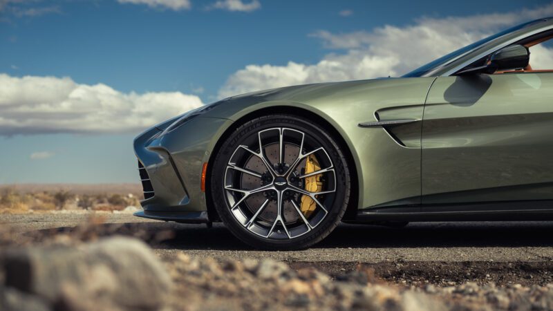 Close-up of the front side of a green sports car parked outdoors, showing the wheel, tire, yellow brake caliper, and part of the fender against a background of sky and clouds.