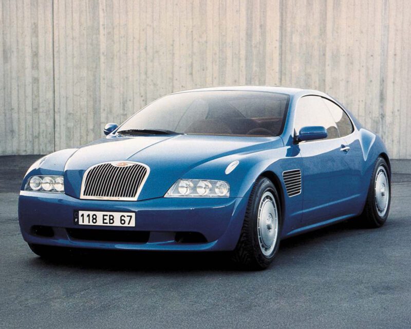 A blue four-door luxury sedan is parked on a paved surface in front of a concrete wall.