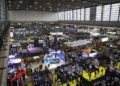 Large indoor car exhibition with numerous booths, display vehicles, and crowds of attendees, reminiscent of the vibrant atmosphere at Rétromobile or a prestigious Auction House like Gooding Christie's, in a spacious, well-lit convention hall.
