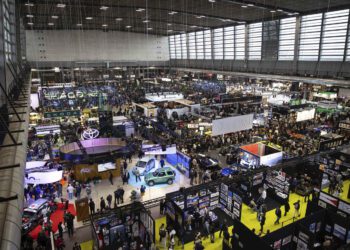 Large indoor car exhibition with numerous booths, display vehicles, and crowds of attendees, reminiscent of the vibrant atmosphere at Rétromobile or a prestigious Auction House like Gooding Christie's, in a spacious, well-lit convention hall.