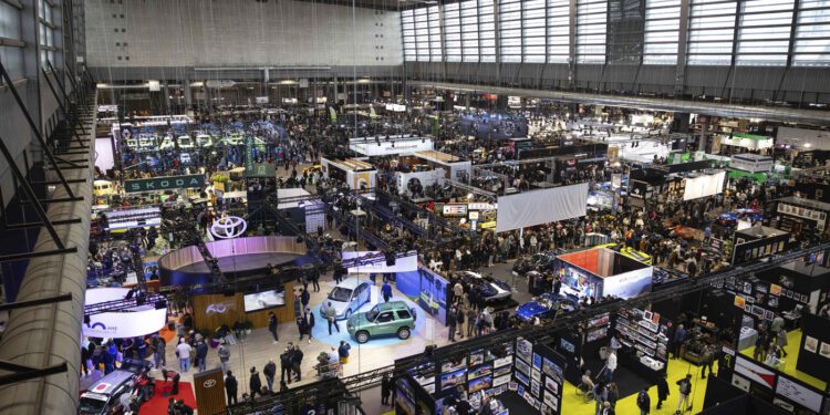 Large indoor car exhibition with numerous booths, display vehicles, and crowds of attendees, reminiscent of the vibrant atmosphere at Rétromobile or a prestigious Auction House like Gooding Christie's, in a spacious, well-lit convention hall.