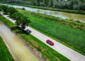 A red Ferrari convertible from the Cavalcade Siviglia drives along a rural road in Andalusia, bordered by a canal and river, surrounded by green fields and trees.