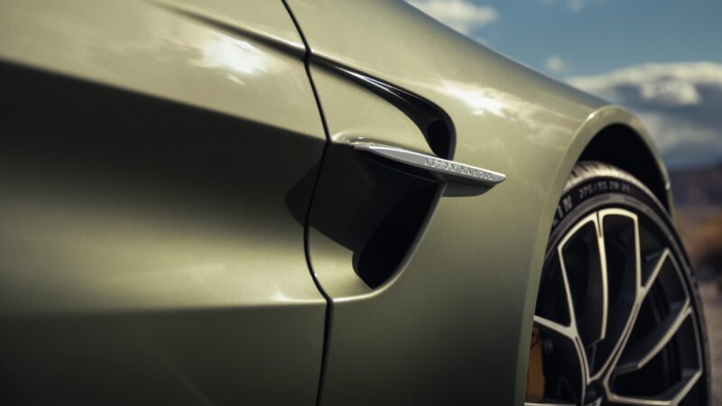 Close-up view of the side vent and front wheel of an Aston Martin car, with badge visible and background slightly blurred.