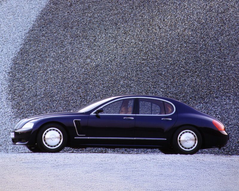 A black luxury sedan is parked in front of a textured, gray stone wall, shown in a profile view with all four doors and wheels visible.
