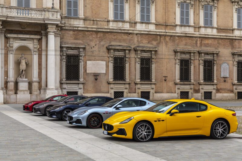 Five Maserati GranTurismo sports cars in different colors are parked in a row in front of a historic building with large windows and stone statues, showcasing the spirit of Modena.