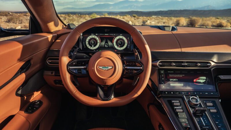 Close-up view of a luxury car's cockpit, featuring a brown leather steering wheel, digital instrument cluster, and central touchscreen display with various controls.