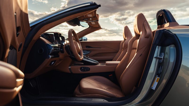 Interior view of a luxury convertible car with tan leather seats, dashboard, and steering wheel, shown with the top down under an open sky.