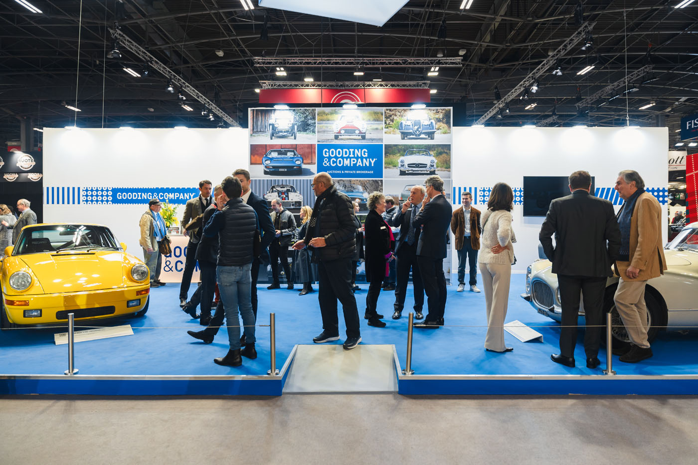 A group of people stand and converse around classic cars at a Gooding & Company exhibition booth during the Rétromobile auction in a convention center.