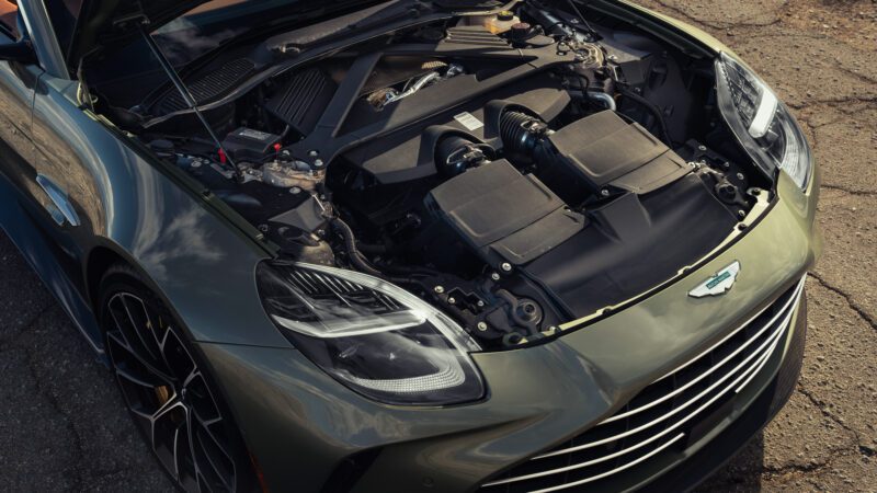 A close-up view of a luxury car’s engine bay with the hood open, showing engine components and the front grille; the car is parked on a rough surface.
