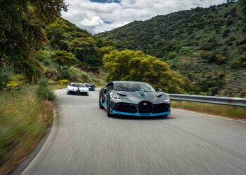 Three sports cars drive on a winding mountain road surrounded by greenery and hills under a partly cloudy sky.
