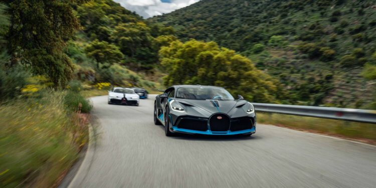 Three sports cars drive on a winding mountain road surrounded by greenery and hills under a partly cloudy sky.