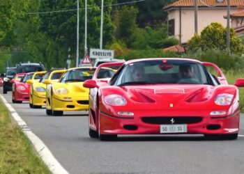 A line of red and yellow Ferrari F50 sports cars drives down a rural road bordered by grassy fields and houses, celebrating the Legacy Tour’s 30th Anniversary.