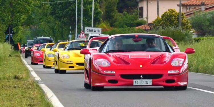 A line of red and yellow Ferrari F50 sports cars drives down a rural road bordered by grassy fields and houses, celebrating the Legacy Tour’s 30th Anniversary.