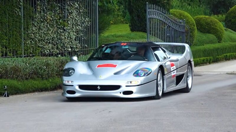 A silver Ferrari F50 sports car is driving on a paved road beside a manicured hedge and an ornate wrought iron gate, capturing the spirit of the Legacy Tour's 30th Anniversary celebration.