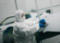 Person in full protective suit and gloves uses a sanding tool on a Modena car hood in a workshop, with other vehicles visible in the background.