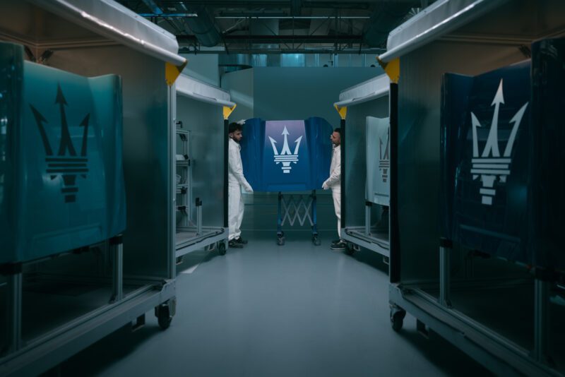Two people in white coveralls stand on either side of a large blue panel featuring the Maserati trident logo, inside Modena’s industrial facility where GranTurismo and GranCabrio models come to life among metal carts.