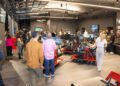 People are gathered in a modern Houston showroom with racing simulators, large screens, and display shelves under bright lighting at the RAFA Racing Club.