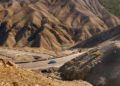 A blue Ferrari drives on a winding road through a dry, mountainous landscape in Andalusia, with brown, rugged hills in the background.