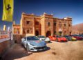 A row of luxury sports cars, including Ferraris, lines up before a rustic stone building in sun-soaked Andalusia under a clear blue sky—a perfect scene from the Cavalcade Siviglia.