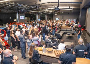 A large group gathers indoors, some seated and others standing, watching a speaker address the crowd in a modern, automotive-themed Houston event space hosted by RAFA Racing Club.