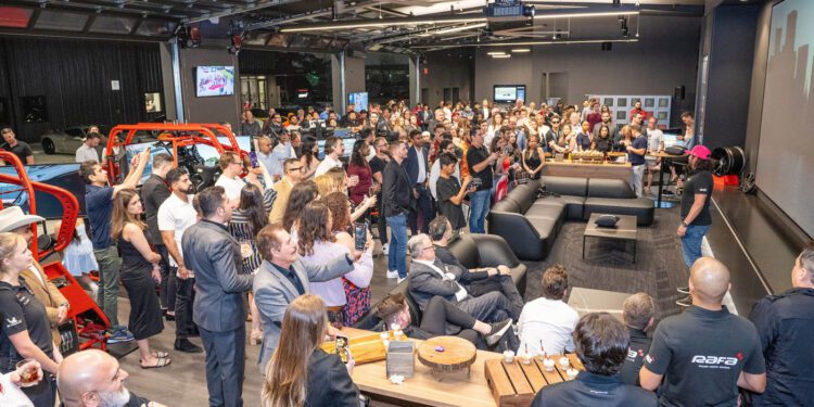 A large group gathers indoors, some seated and others standing, watching a speaker address the crowd in a modern, automotive-themed Houston event space hosted by RAFA Racing Club.