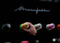 A hand selects a spool of yellow thread from a display of colorful thread spools mounted on a black board labeled "Manufaktur," reminiscent of the precision and luxury found in Mercedes-Benz gifts.