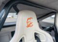 Close-up of a white car seat with orange stitching and an orange scorpion logo, featuring a roll cage and harness, inside a modern 1,000-HP Hellephant-powered sports car inspired by the 1969 Charger.
