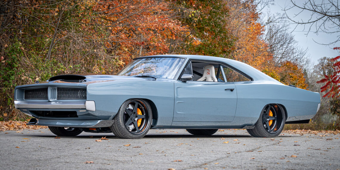 A light blue 1969 Charger classic muscle car with black wheels, yellow brake calipers, and a hood scoop is parked on a paved road with autumn foliage in the background—a true Hellephant-powered icon.