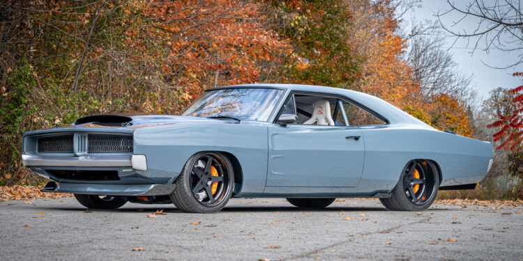 A light blue 1969 Charger classic muscle car with black wheels, yellow brake calipers, and a hood scoop is parked on a paved road with autumn foliage in the background—a true Hellephant-powered icon.