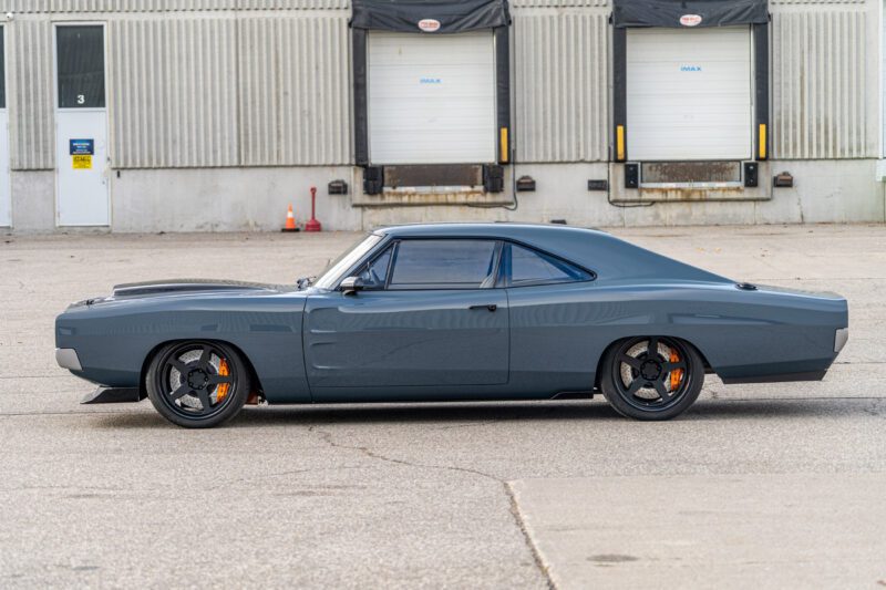 Side view of a gray 1969 Charger muscle car with black wheels, featuring a powerful Hellephant engine, parked in front of a warehouse loading dock.
