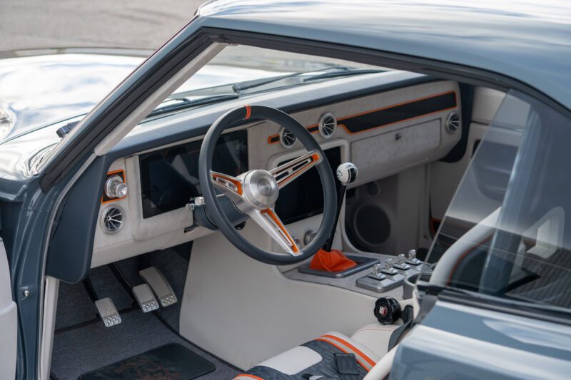 The interior of a modernized 1969 Charger Hellephant features a minimalist dashboard, metal steering wheel, three pedals, and custom controls with striking orange accents.
