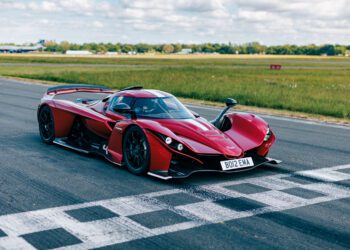 A red supercar is parked on the Top Gear Test Track at the starting line, with grass and trees in the background under a partly cloudy sky, ready to chase a new lap record.
