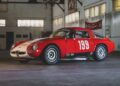 A classic red and white race car with the number 199 on the side is parked inside an industrial garage with large windows, set to be featured at the RM Sotheby’s Quadrifoglio Collection Monterey 2025.