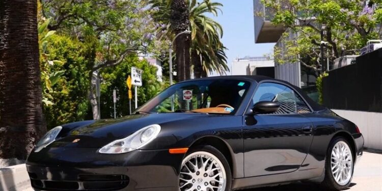 A black Porsche 911 convertible, one of the best 2000s cars, is parked on a sunny street lined with palm trees and modern buildings. A phone number for cars for sale appears at the bottom right of the image.