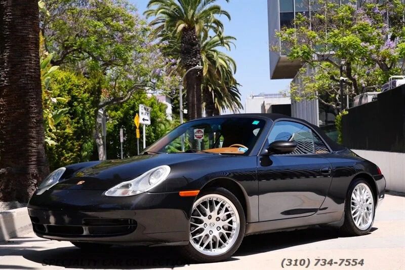 A black Porsche 911 convertible, one of the best 2000s cars, is parked on a sunny street lined with palm trees and modern buildings. A phone number for cars for sale appears at the bottom right of the image.