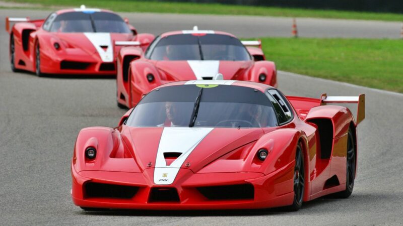 Three red Ferrari FXX sports cars with white racing stripes, featured among the 10 Enzo Derivates That Only REAL Ferrari Fans Know About, are speeding on a racetrack bordered by green grass.