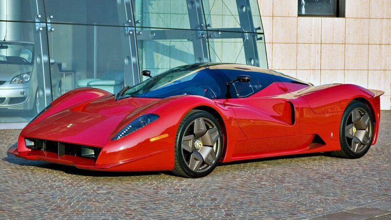 A red Ferrari P4/5 sports car, one of the 10 Enzo Derivates That Only REAL Ferrari Fans Know About, is parked on a cobblestone surface in front of a modern glass building.