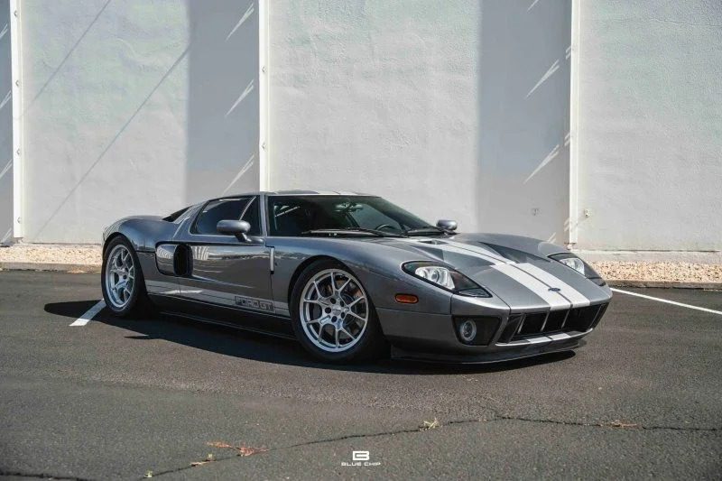 A silver Ford GT sports car with white racing stripes, one of the best 2000s cars, is parked on a paved lot beside a white wall.