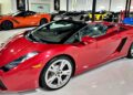 A red Lamborghini Gallardo Spyder convertible is parked indoors among several other luxury sports cars in a showroom, showcasing why it’s considered one of the Best Used Cars For $100000.