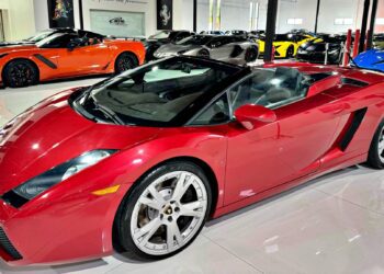 A red Lamborghini Gallardo Spyder convertible is parked indoors among several other luxury sports cars in a showroom, showcasing why it’s considered one of the Best Used Cars For $100000.