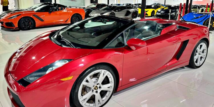 A red Lamborghini Gallardo Spyder convertible is parked indoors among several other luxury sports cars in a showroom, showcasing why it’s considered one of the Best Used Cars For $100000.