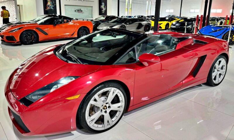A red Lamborghini Gallardo Spyder convertible is parked indoors among several other luxury sports cars in a showroom, showcasing why it’s considered one of the Best Used Cars For $100000.