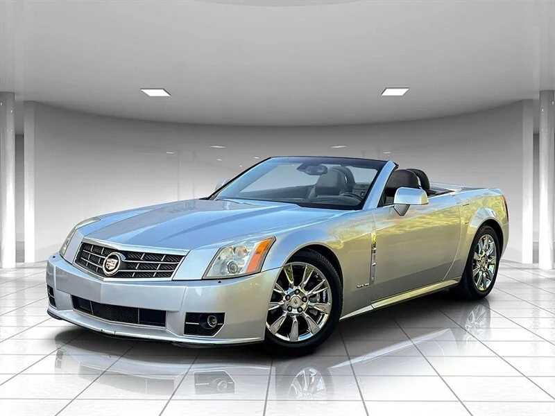 Silver convertible sports car, one of the best 2000s cars, viewed from the front left, parked indoors on a reflective tiled floor with a curved wall and ceiling lights above.