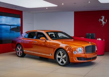An orange Bentley Mulsanne, one of the best Bentleys, is displayed in a modern showroom with red walls and a Ferrari logo in the background.