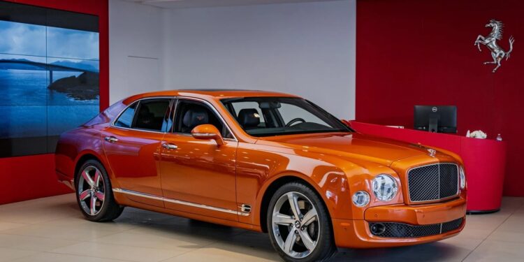 An orange Bentley Mulsanne, one of the best Bentleys, is displayed in a modern showroom with red walls and a Ferrari logo in the background.