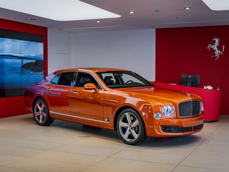 An orange Bentley Mulsanne, one of the best Bentleys, is displayed in a modern showroom with red walls and a Ferrari logo in the background.